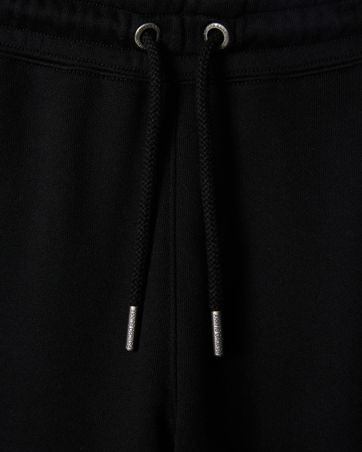 Close-up of black sweatpants, known as the KIDS PACIFIC CHENILLE JOGGER - B0P972D200 by Psycho Bunny, featuring a ribbed elastic waistband with a black drawstring. The drawstring has metallic tips, and the fabric appears soft and comfortable. A high-pile chenille logo patch adds a stylish touch to this casual bottom.