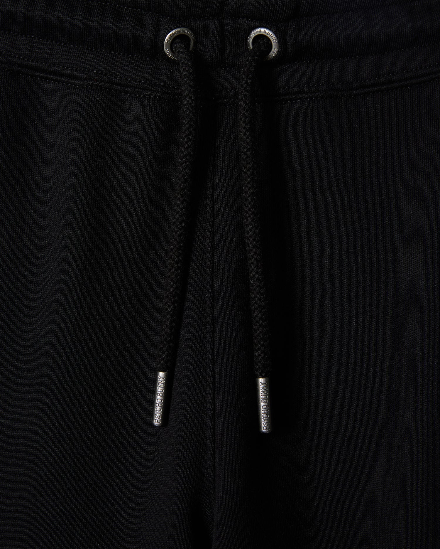 Close-up of black sweatpants, known as the KIDS PACIFIC CHENILLE JOGGER - B0P972D200 by Psycho Bunny, featuring a ribbed elastic waistband with a black drawstring. The drawstring has metallic tips, and the fabric appears soft and comfortable. A high-pile chenille logo patch adds a stylish touch to this casual bottom.