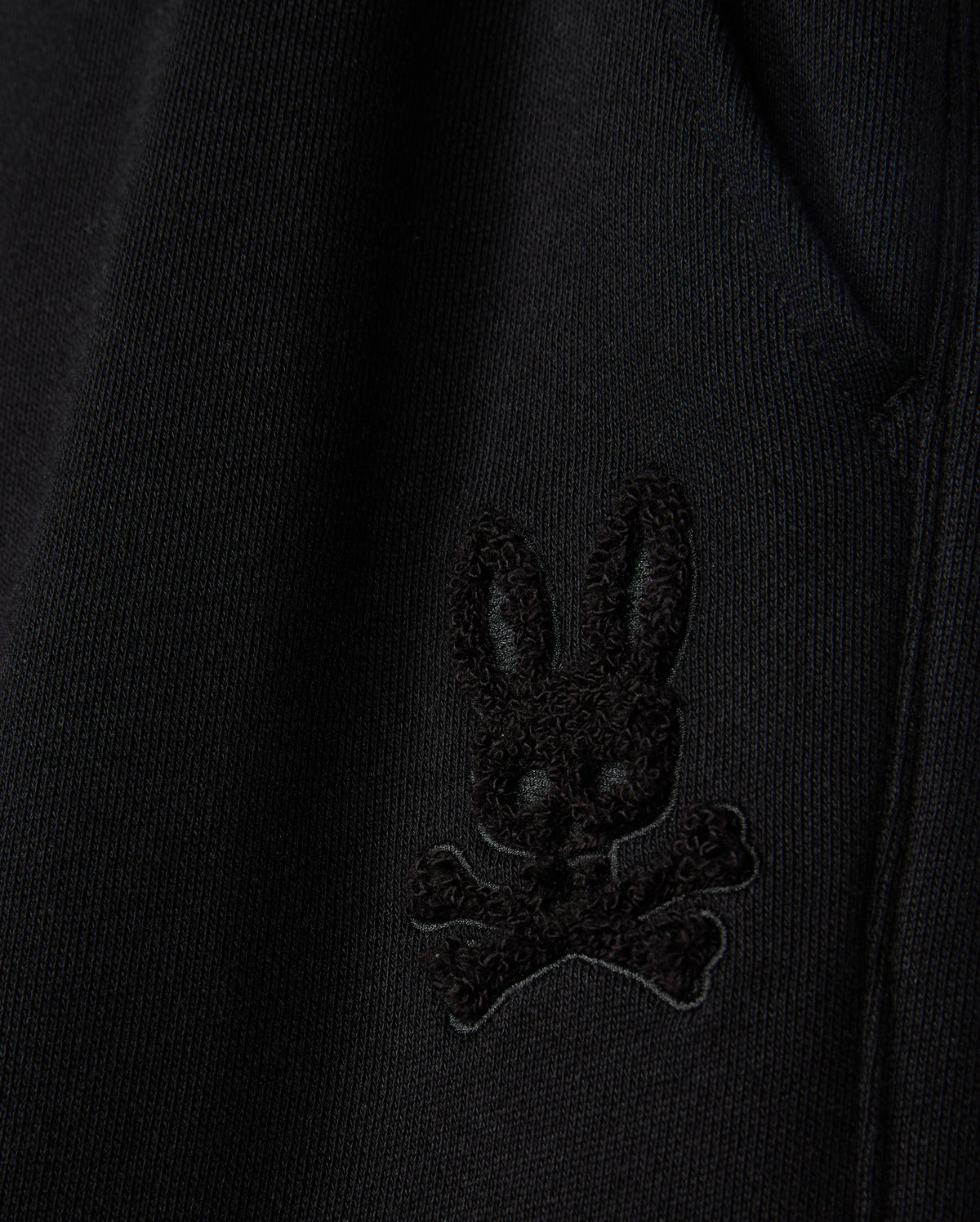 Close-up of the fabric from the KIDS PACIFIC CHENILLE JOGGER by Psycho Bunny, featuring an embroidered patch with a stylized black bunny head above crossed bones. This high-pile chenille logo patch adds a distinctive touch to any casual bottom or jogger pant.