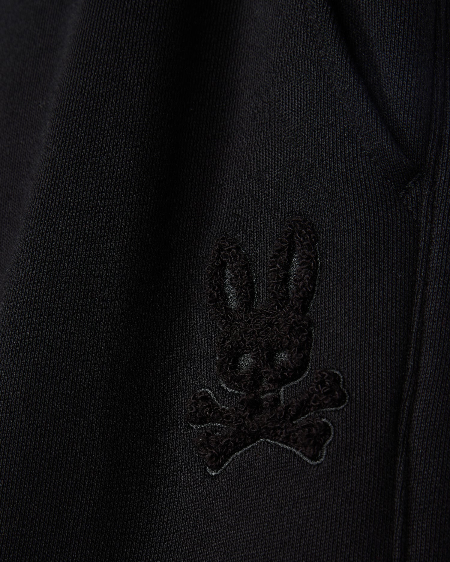 Close-up of the fabric from the KIDS PACIFIC CHENILLE JOGGER by Psycho Bunny, featuring an embroidered patch with a stylized black bunny head above crossed bones. This high-pile chenille logo patch adds a distinctive touch to any casual bottom or jogger pant.