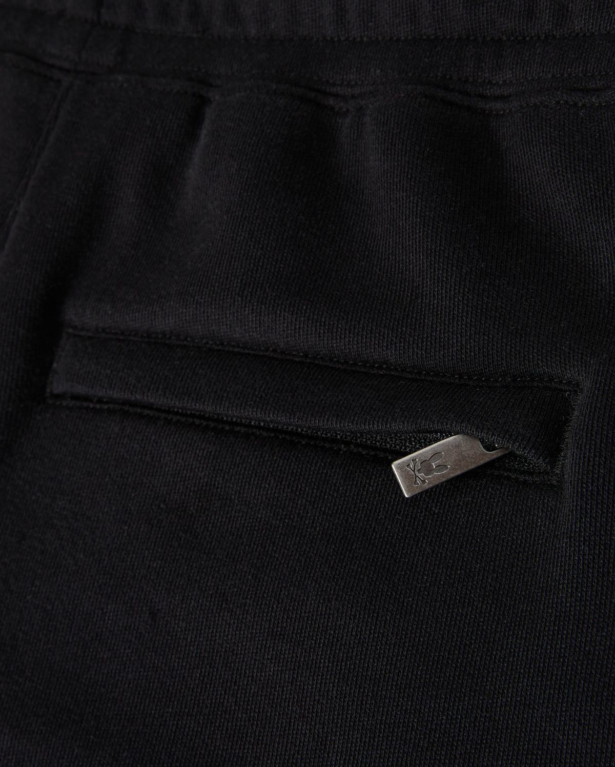 Close-up of a black fabric garment, possibly the Kids Pacific Chenille Jogger by Psycho Bunny, featuring a pocket with a horizontal zipper. The metallic zipper pull includes a small design or logo. The texture of the fabric appears soft and smooth, enhancing its casual bottom style.