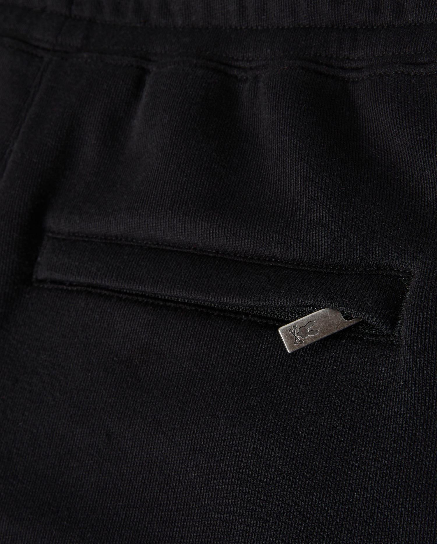 Close-up of a black fabric garment, possibly the Kids Pacific Chenille Jogger by Psycho Bunny, featuring a pocket with a horizontal zipper. The metallic zipper pull includes a small design or logo. The texture of the fabric appears soft and smooth, enhancing its casual bottom style.