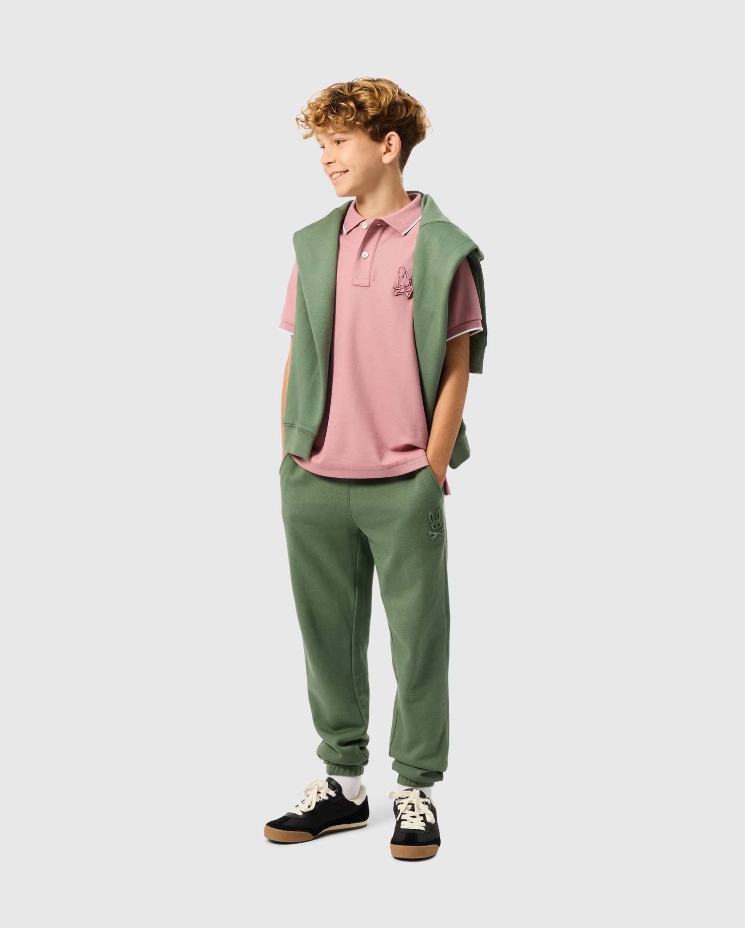 A young boy with curly hair stands smiling, wearing a pink polo shirt and green joggers adorned with the Psycho Bunny logo. He finishes his look with black sneakers and has a matching green sweatshirt draped over his shoulders. The background is plain white.