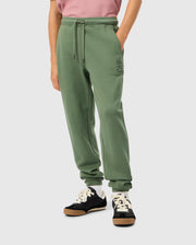A person is wearing the Psycho Bunny KIDS PACIFIC CHENILLE JOGGER - B0P972D200, which features a chenille logo patch on the left thigh, paired with a pink top and black sneakers with white laces. Their hands rest casually in their jogger pockets.