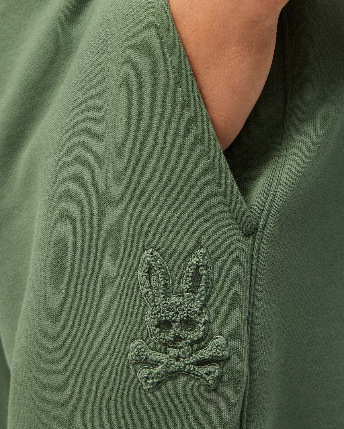 Close-up of the KIDS PACIFIC CHENILLE JOGGER in green by Psycho Bunny, showcasing an embroidered patch depicting a playful bunny head with crossed bones underneath. The chenille logo patch adds texture and enhances the unique charm of this casual bottom.