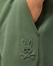 Close-up of the KIDS PACIFIC CHENILLE JOGGER in green by Psycho Bunny, showcasing an embroidered patch depicting a playful bunny head with crossed bones underneath. The chenille logo patch adds texture and enhances the unique charm of this casual bottom.