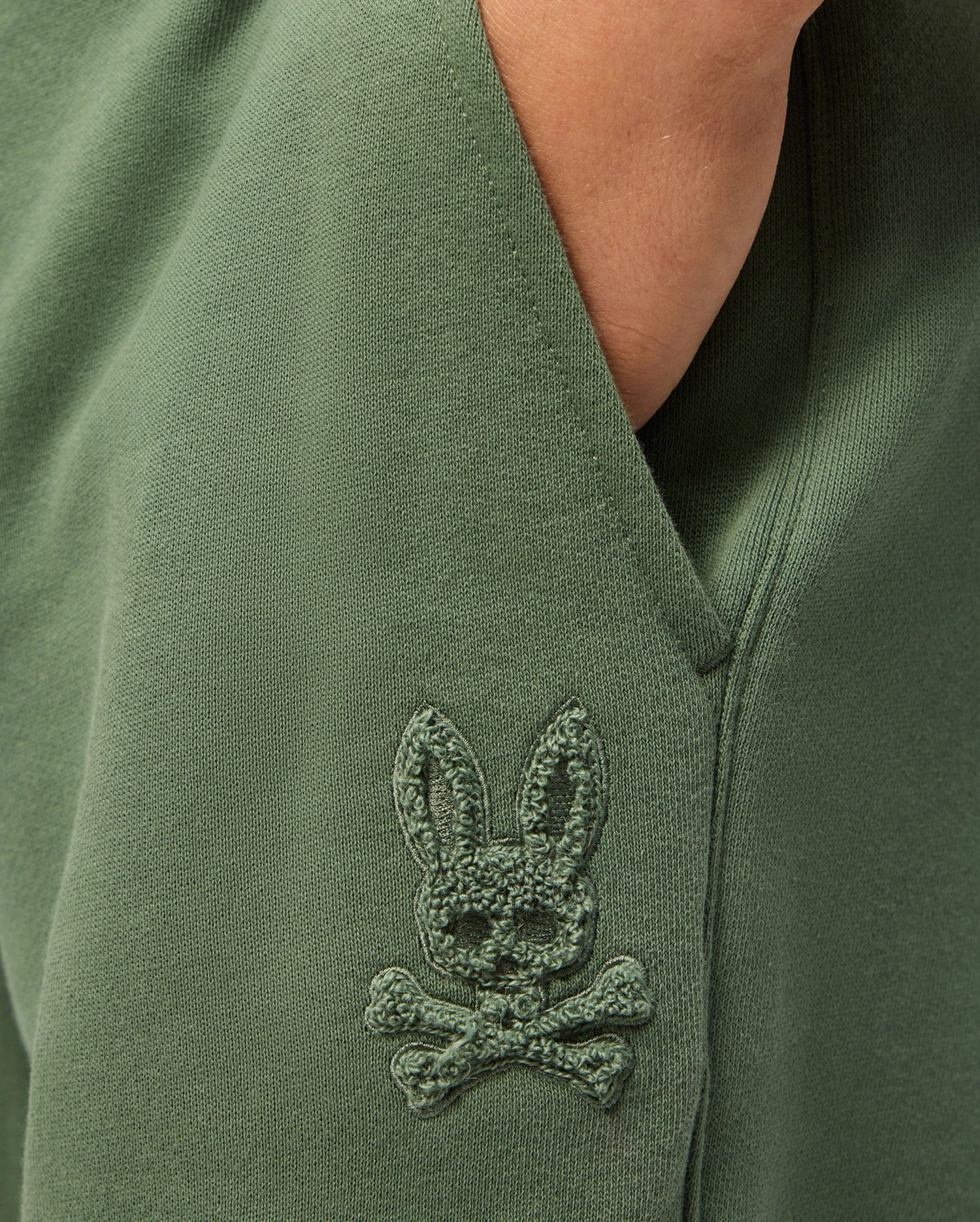 Close-up of the KIDS PACIFIC CHENILLE JOGGER in green by Psycho Bunny, showcasing an embroidered patch depicting a playful bunny head with crossed bones underneath. The chenille logo patch adds texture and enhances the unique charm of this casual bottom.
