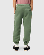 Wearing the KIDS PACIFIC CHENILLE JOGGER - B0P972D200 from Psycho Bunny, a person casually dressed in green faces away with hands in their pockets. These joggers feature a distinctive chenille logo patch and a right-side back pocket. They complete their look with black sneakers featuring white soles, perfectly paired with a pink shirt.