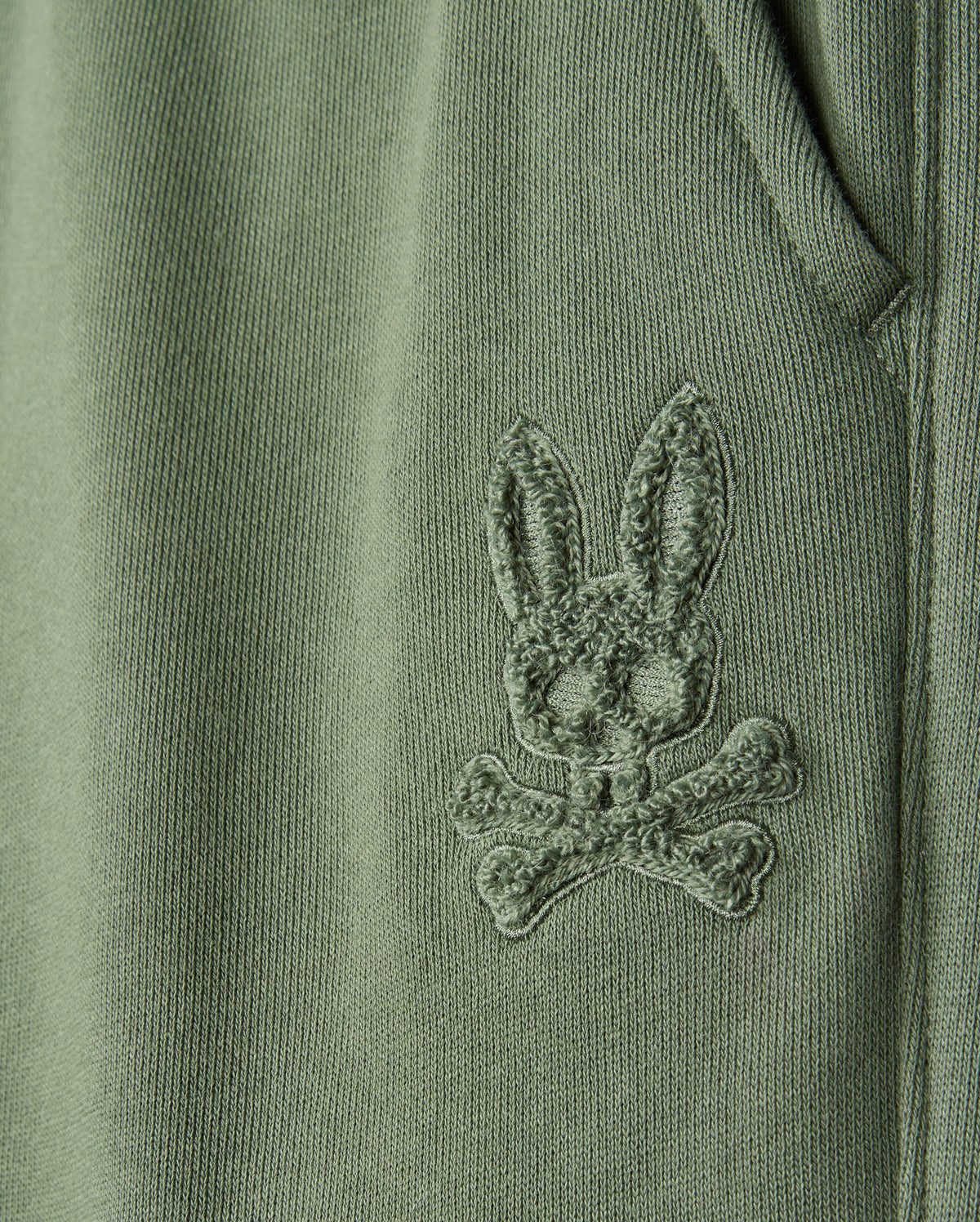 Close-up of a green fabric from the KIDS PACIFIC CHENILLE JOGGER - B0P972D200 by Psycho Bunny, featuring an embroidered bunny's head with crossbones beneath it, reminiscent of a chenille logo patch. The detailed stitching adds a textured appearance to the material.