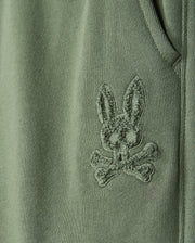 Close-up of a green fabric from the KIDS PACIFIC CHENILLE JOGGER - B0P972D200 by Psycho Bunny, featuring an embroidered bunny's head with crossbones beneath it, reminiscent of a chenille logo patch. The detailed stitching adds a textured appearance to the material.
