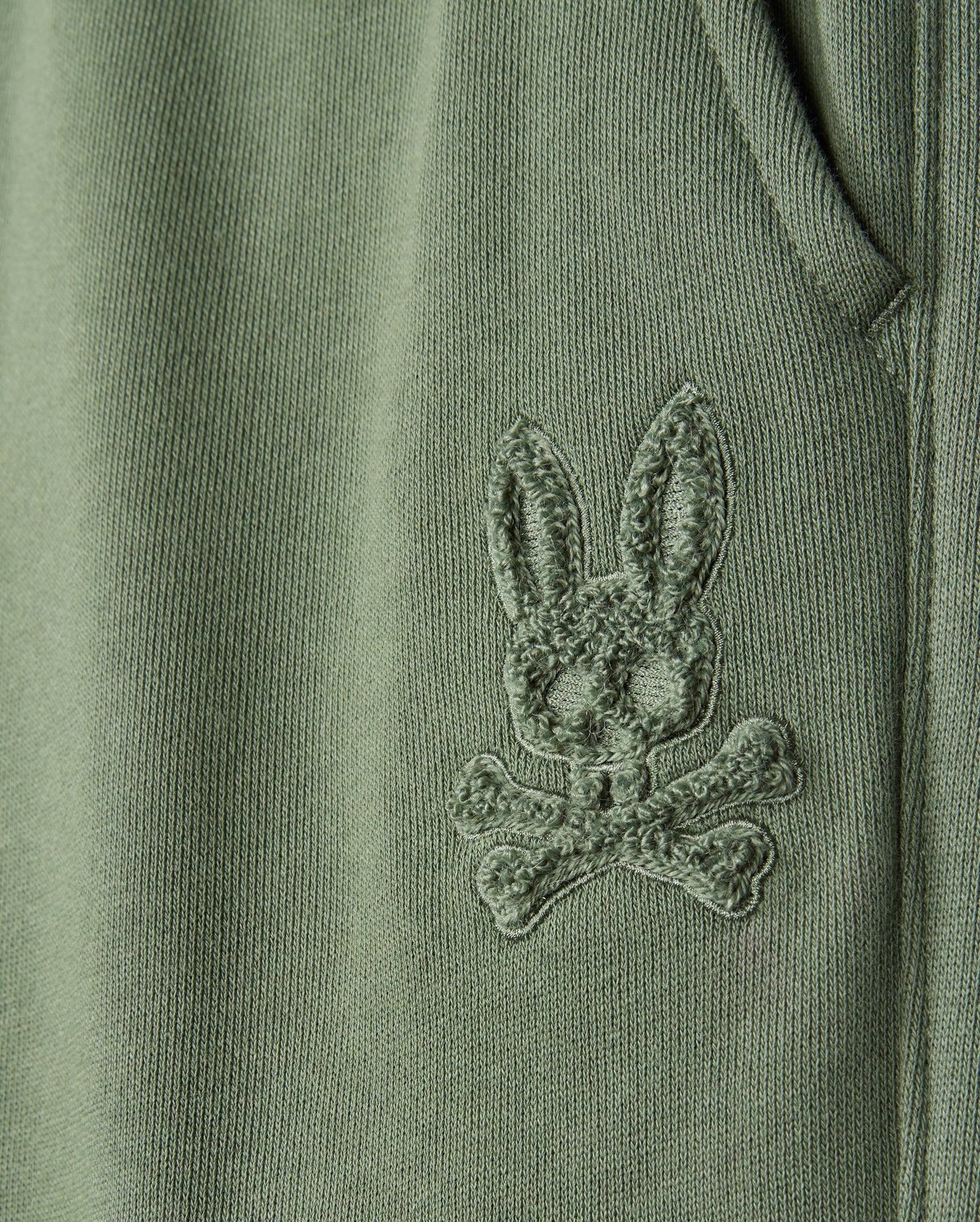 Close-up of a green fabric from the KIDS PACIFIC CHENILLE JOGGER - B0P972D200 by Psycho Bunny, featuring an embroidered bunny's head with crossbones beneath it, reminiscent of a chenille logo patch. The detailed stitching adds a textured appearance to the material.