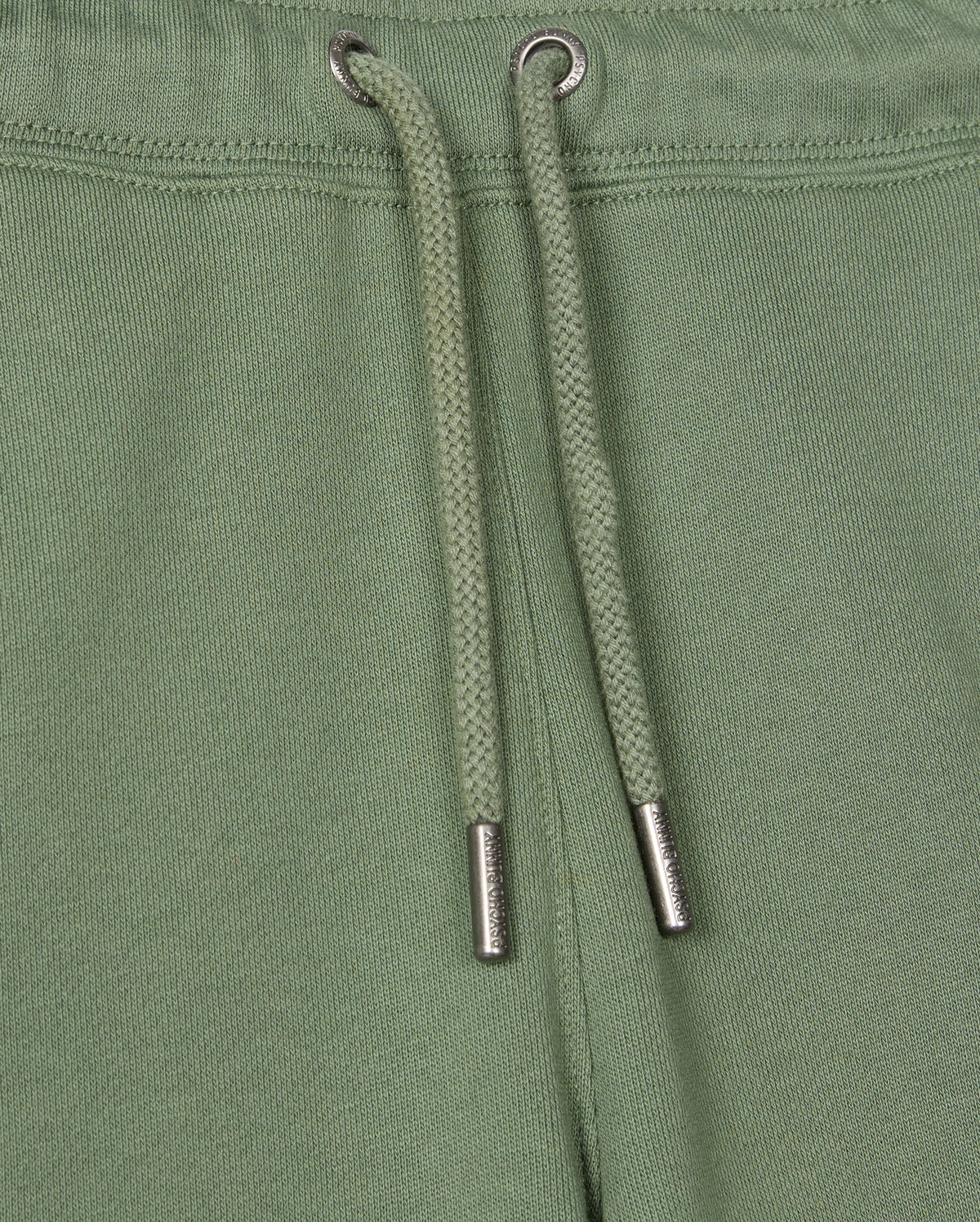 Close-up of the Kids Pacific Chenille Jogger from Psycho Bunny, showcasing green jogger pants with a drawstring featuring metal tips. These casual bottoms are made with textured fabric and visible stitching near the top edge, enhanced by a chenille logo patch for added style.