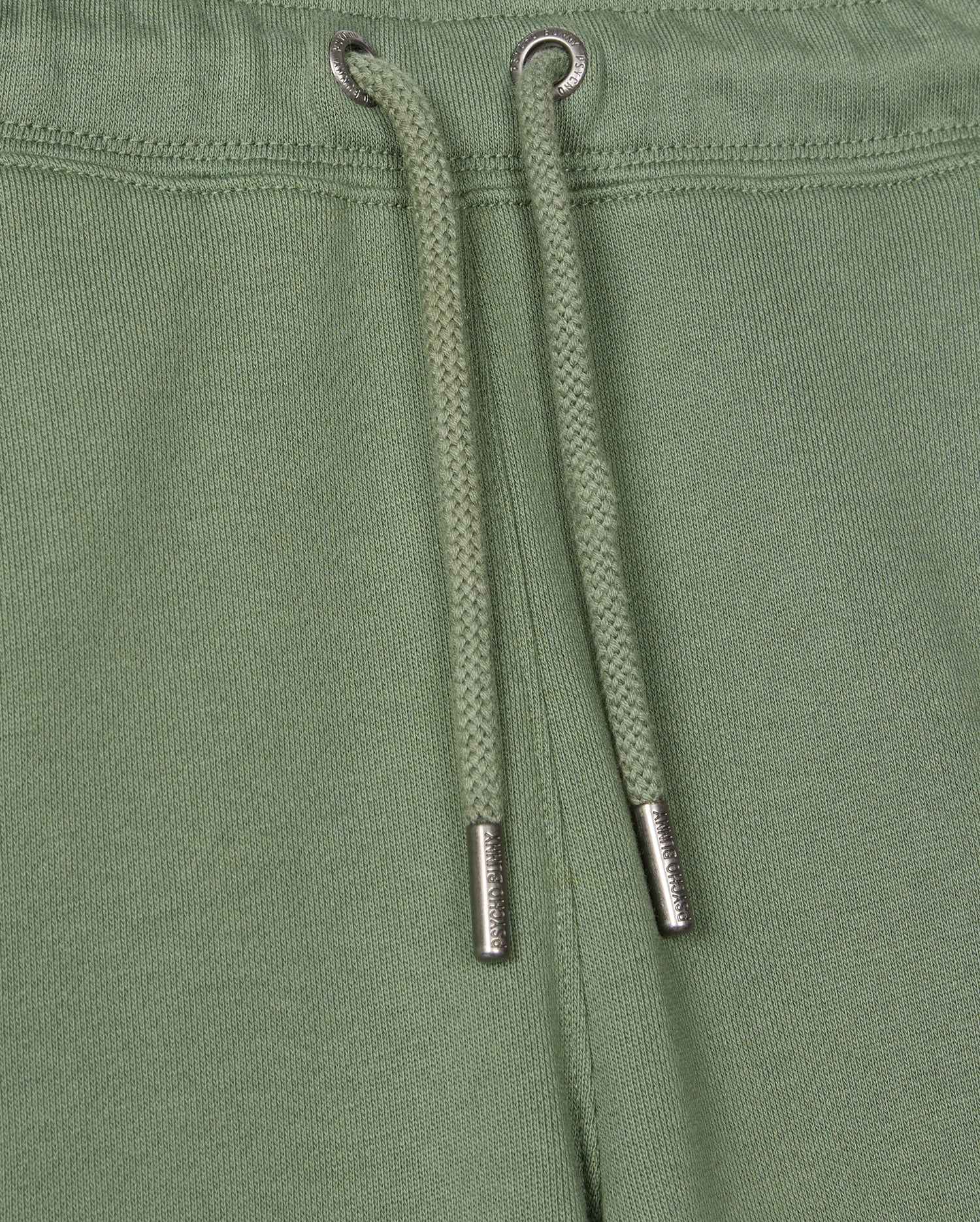 Close-up of the Kids Pacific Chenille Jogger from Psycho Bunny, showcasing green jogger pants with a drawstring featuring metal tips. These casual bottoms are made with textured fabric and visible stitching near the top edge, enhanced by a chenille logo patch for added style.