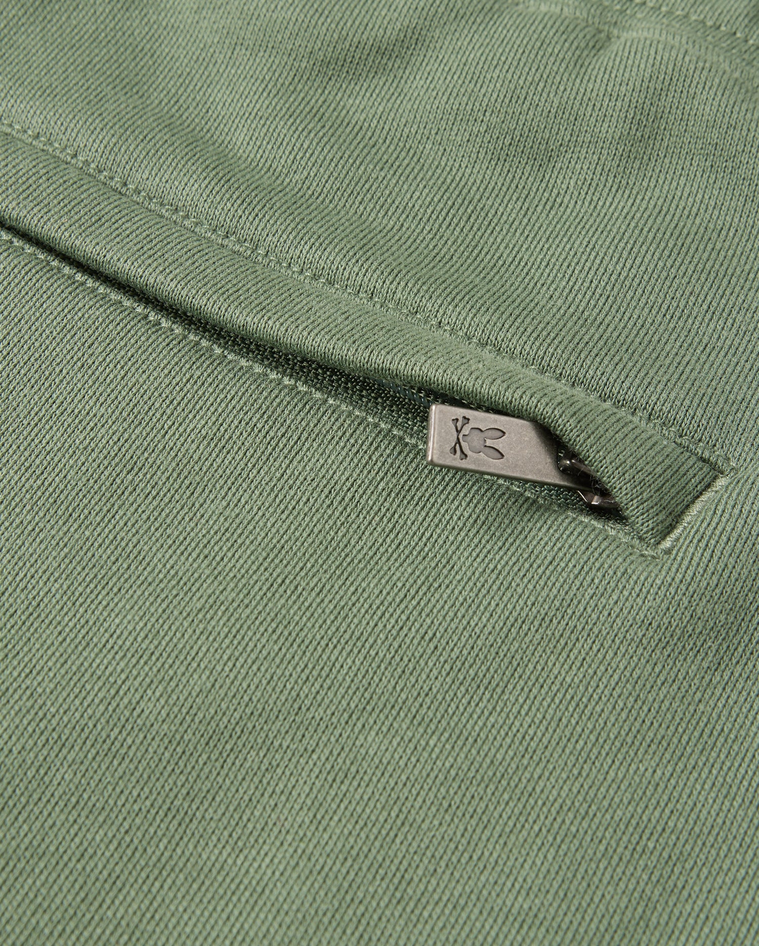 Close-up of the green **KIDS PACIFIC CHENILLE JOGGER - B0P972D200** by **Psycho Bunny**, featuring a subtle metal zipper detail. The zipper, adorned with a small decorative symbol on its pull tab, complements the jogger's textured woven pattern and casual bottom style.