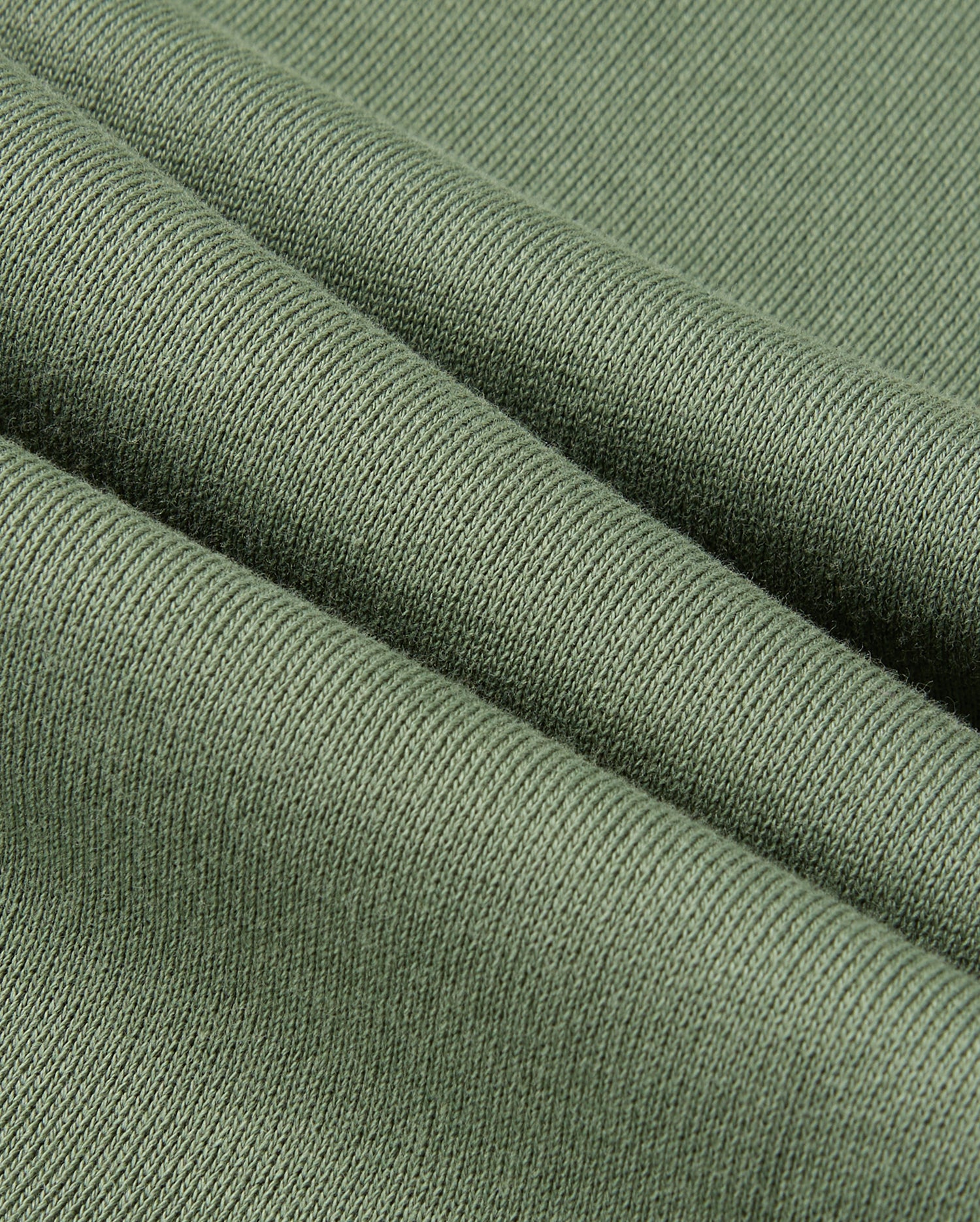 A close-up of a textured green fabric folded in layers, reminiscent of the KIDS PACIFIC CHENILLE JOGGER by Psycho Bunny. The ribbed pattern highlights its softness and thickness, with gentle diagonal folds creating shadows and highlights similar to those found in a jogger pant.