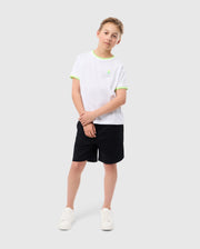 A young boy stands against a plain background, wearing a slim fit white T-shirt with light green accents at the sleeves and collar, paired with black shorts and white sneakers. The Psycho Bunny KIDS WILLIS STRETCH TENCEL SHORT - B0R239Y1WB ensures comfort. He has short, light brown hair and is posing casually with one leg slightly bent.