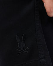 Close-up of KIDS WILLIS STRETCH TENCEL SHORT - B0R239Y1WB by Psycho Bunny featuring an embroidered Bunny logo with crossed bones underneath. A person's hand is visible at the top right corner, partially gripping the fabric. The background is out of focus.