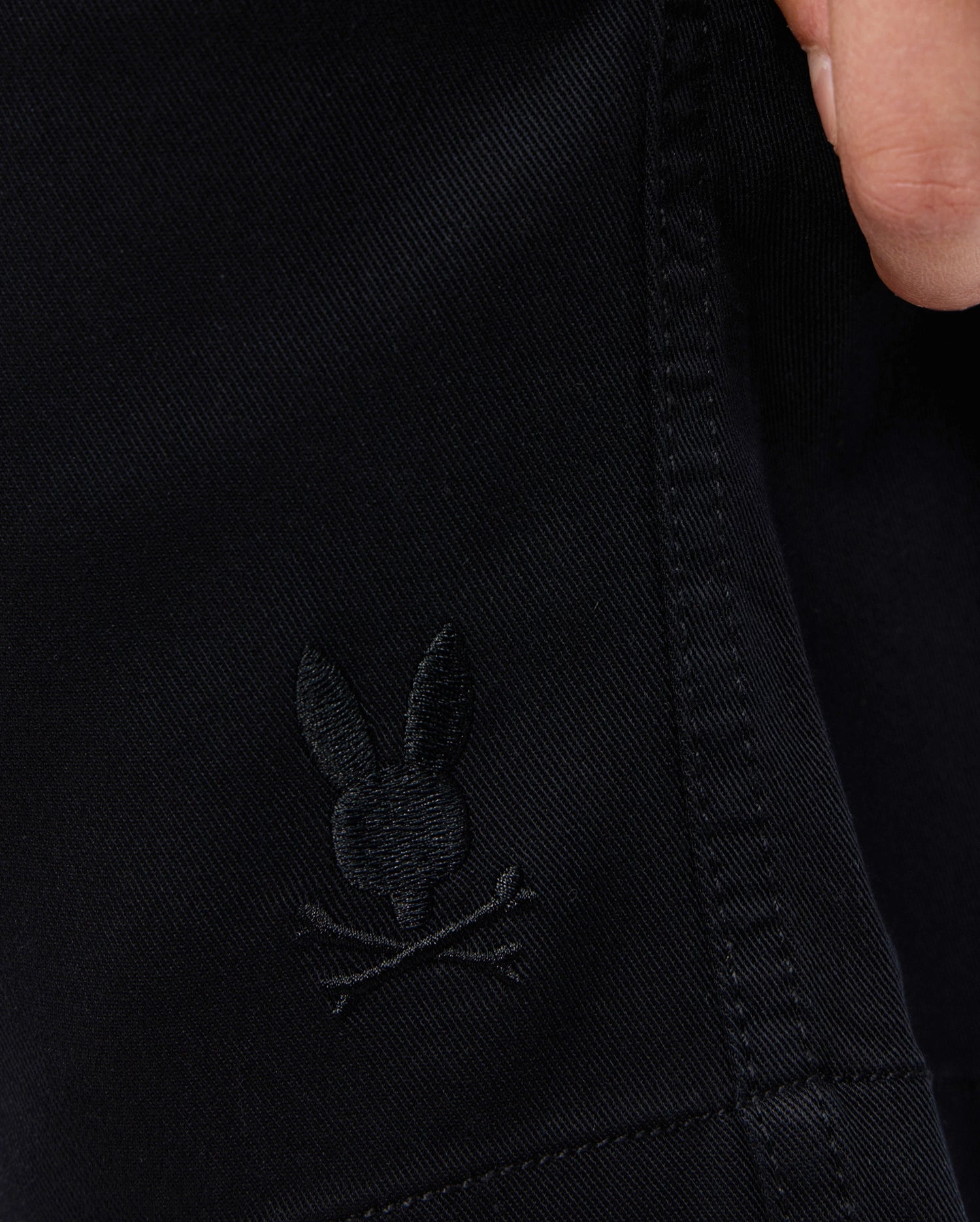 Close-up of KIDS WILLIS STRETCH TENCEL SHORT - B0R239Y1WB by Psycho Bunny featuring an embroidered Bunny logo with crossed bones underneath. A person's hand is visible at the top right corner, partially gripping the fabric. The background is out of focus.
