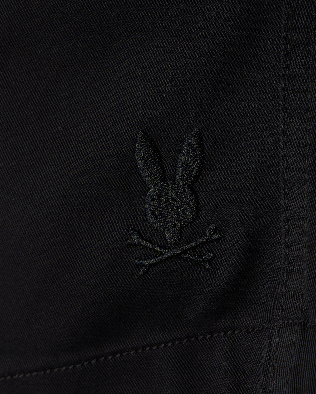 Close-up of black **KIDS WILLIS STRETCH TENCEL SHORT - B0R239Y1WB** fabric featuring an embroidered Bunny logo with ears upright, situated above a pair of crossed bones, reminiscent of a skull and crossbones motif. The slim fit fabric has visible stitching details. **Brand Name: Psycho Bunny**