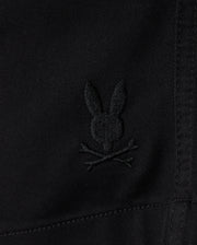 Close-up of black **KIDS WILLIS STRETCH TENCEL SHORT - B0R239Y1WB** fabric featuring an embroidered Bunny logo with ears upright, situated above a pair of crossed bones, reminiscent of a skull and crossbones motif. The slim fit fabric has visible stitching details. **Brand Name: Psycho Bunny**