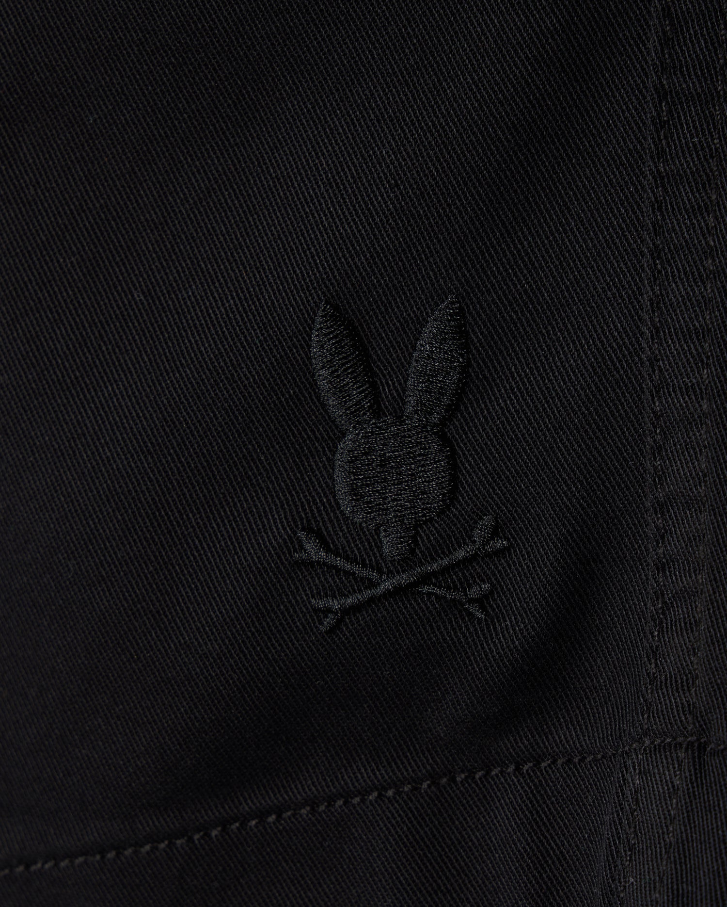 Close-up of black **KIDS WILLIS STRETCH TENCEL SHORT - B0R239Y1WB** fabric featuring an embroidered Bunny logo with ears upright, situated above a pair of crossed bones, reminiscent of a skull and crossbones motif. The slim fit fabric has visible stitching details. **Brand Name: Psycho Bunny**