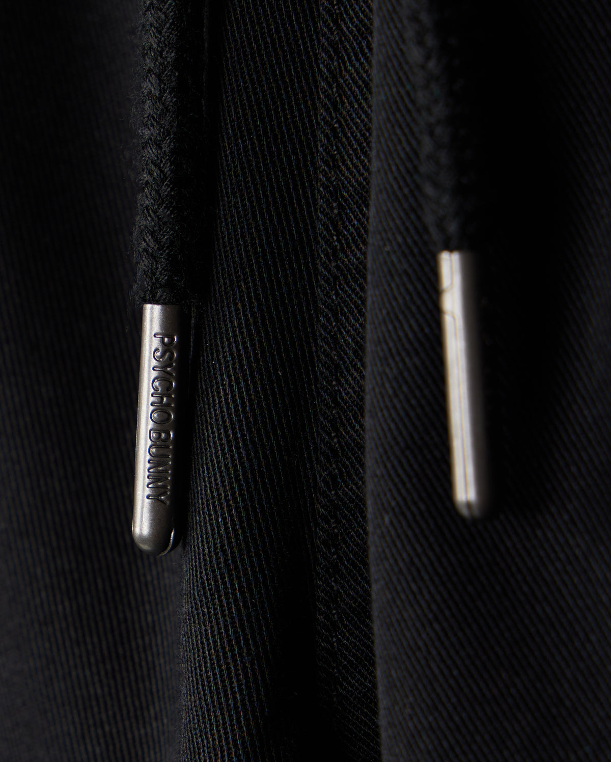 Close-up image of two black drawstrings with metallic tips, one labeled with the words "PSYCHO BUNNY". The background is a black fabric, possibly part of a hoodie or sweatpants featuring an embroidered Bunny logo. The clothing item in question is the KIDS WILLIS STRETCH TENCEL SHORT - B0R239Y1WB by Psycho Bunny.