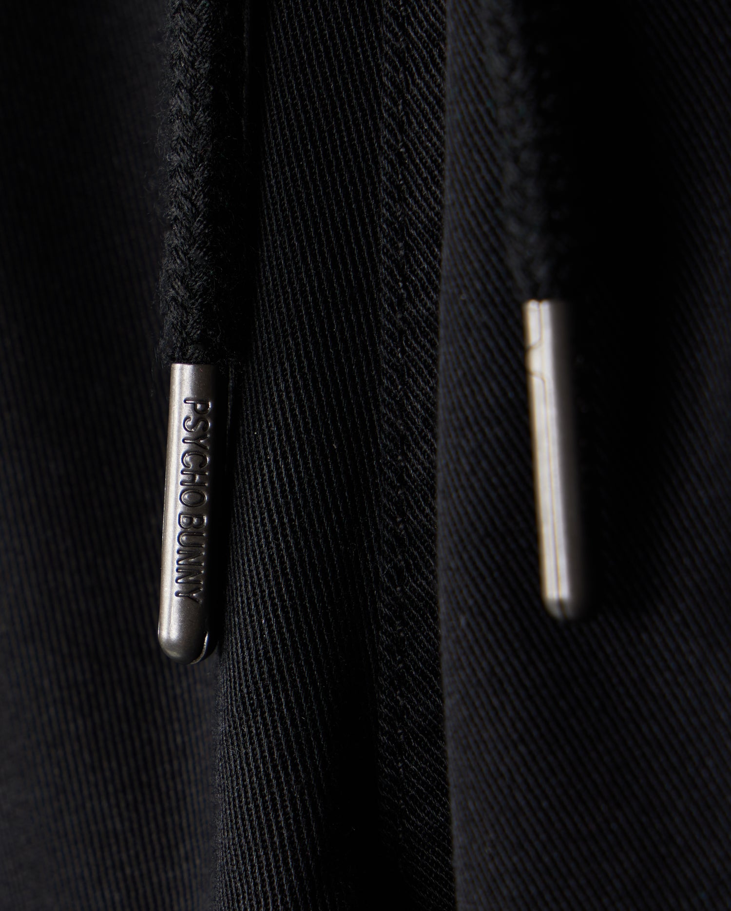 Close-up image of two black drawstrings with metallic tips, one labeled with the words "PSYCHO BUNNY". The background is a black fabric, possibly part of a hoodie or sweatpants featuring an embroidered Bunny logo. The clothing item in question is the KIDS WILLIS STRETCH TENCEL SHORT - B0R239Y1WB by Psycho Bunny.