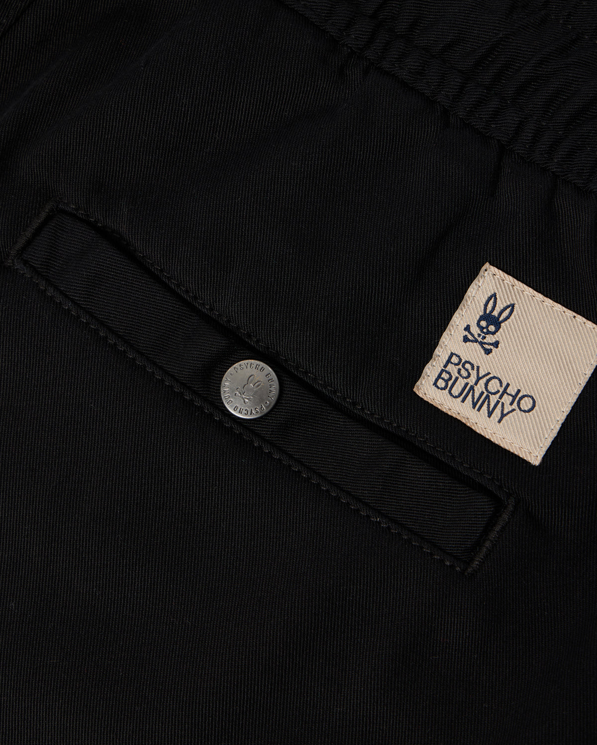 Close-up of black KIDS WILLIS STRETCH TENCEL SHORT - B0R239Y1WB fabric featuring a pocket with a button and a sewn patch label. The label displays the embroidered Bunny logo, consisting of a bunny skull and crossbones, along with the brand name "Psycho Bunny" in text.