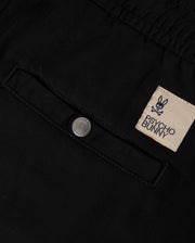 Close-up of black KIDS WILLIS STRETCH TENCEL SHORT - B0R239Y1WB fabric featuring a pocket with a button and a sewn patch label. The label displays the embroidered Bunny logo, consisting of a bunny skull and crossbones, along with the brand name "Psycho Bunny" in text.