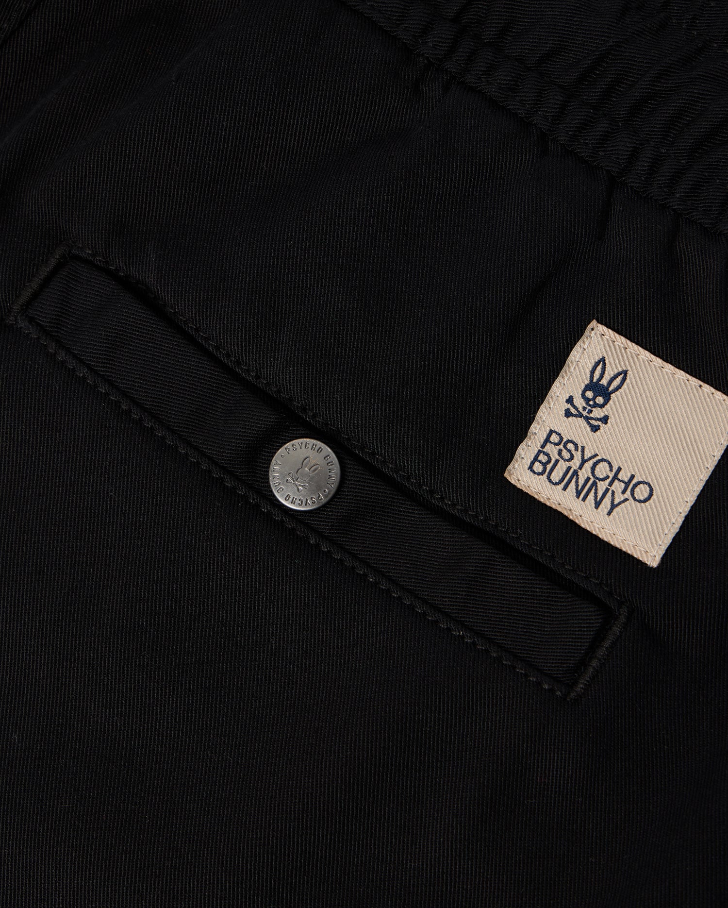 Close-up of black KIDS WILLIS STRETCH TENCEL SHORT - B0R239Y1WB fabric featuring a pocket with a button and a sewn patch label. The label displays the embroidered Bunny logo, consisting of a bunny skull and crossbones, along with the brand name "Psycho Bunny" in text.