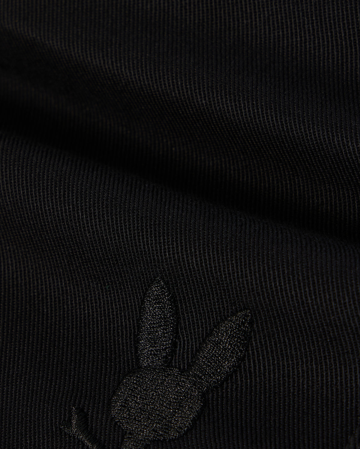 A close-up image of black fabric with a subtle embroidered Bunny logo featuring a rabbit's head with long ears on the lower portion. The texture of the KIDS WILLIS STRETCH TENCEL SHORT - B0R239Y1WB from Psycho Bunny is ribbed, and the embroidery is slightly raised, blending well with the dark color.