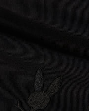 A close-up image of black fabric with a subtle embroidered Bunny logo featuring a rabbit's head with long ears on the lower portion. The texture of the KIDS WILLIS STRETCH TENCEL SHORT - B0R239Y1WB from Psycho Bunny is ribbed, and the embroidery is slightly raised, blending well with the dark color.