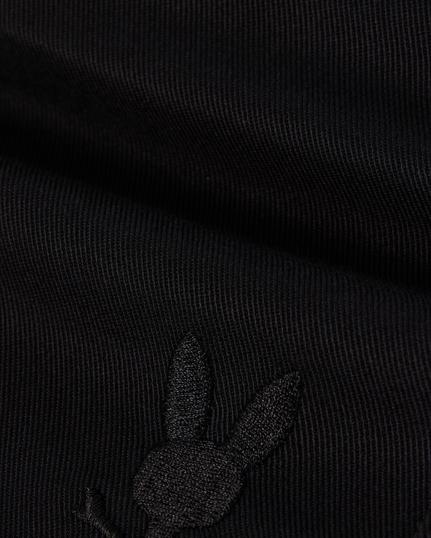 A close-up image of black fabric with a subtle embroidered Bunny logo featuring a rabbit's head with long ears on the lower portion. The texture of the KIDS WILLIS STRETCH TENCEL SHORT - B0R239Y1WB from Psycho Bunny is ribbed, and the embroidery is slightly raised, blending well with the dark color.