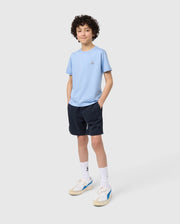 A young boy with curly hair, wearing a light blue t-shirt, Psycho Bunny KIDS WILLIS STRETCH TENCEL SHORT - B0R239Y1WB with an elasticized waistband, white socks, and sneakers, stands smiling against a plain background.