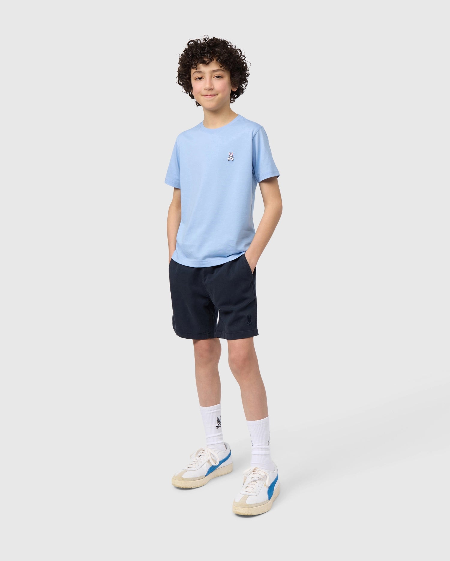 A young boy with curly hair, wearing a light blue t-shirt, Psycho Bunny KIDS WILLIS STRETCH TENCEL SHORT - B0R239Y1WB with an elasticized waistband, white socks, and sneakers, stands smiling against a plain background.