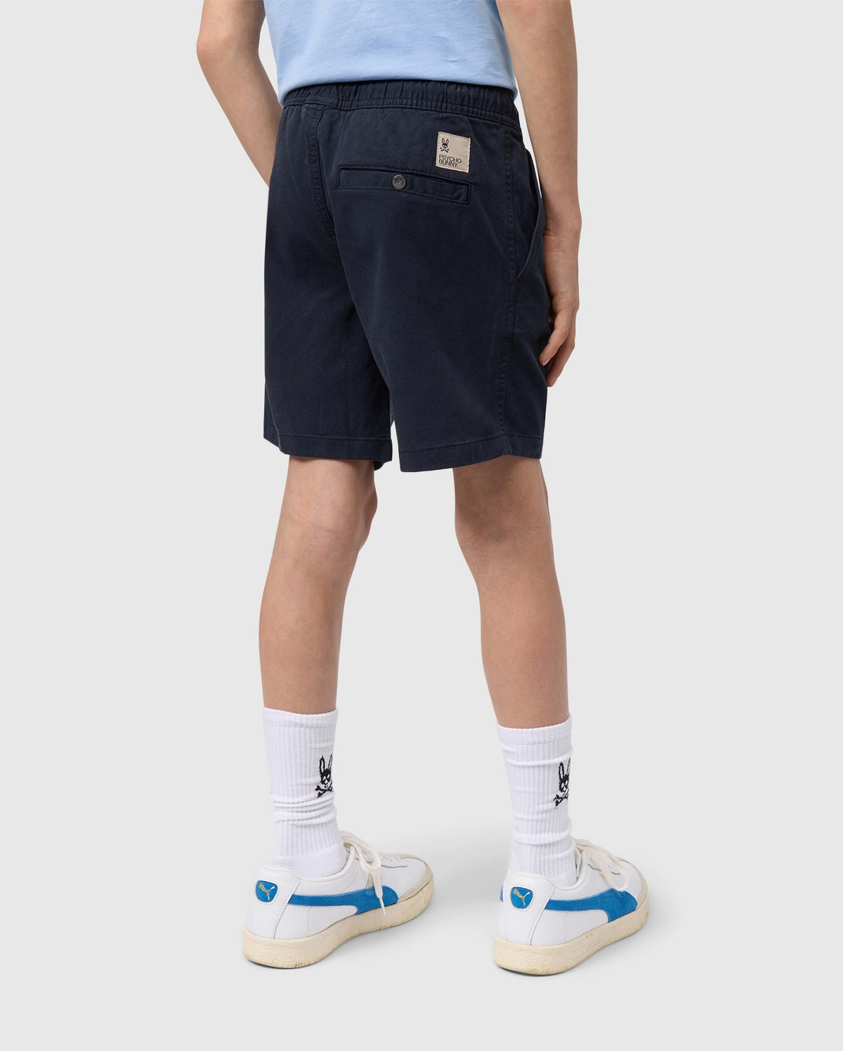 A person from the back wearing navy blue Psycho Bunny kids' shorts, white socks with a logo, and beige sneakers with blue accents. The focus is on the casual, sporty attire.
