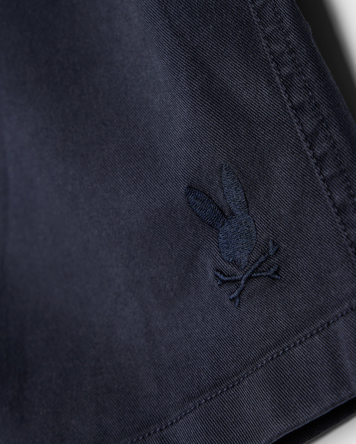 Close-up of navy blue Psycho Bunny kids' shorts crafted from KIDS WILLIS STRETCH TENCEL SHORT - B0R239Y1WB twill with an intricately embroidered logo featuring a rabbit. The texture of the material displays fine threading and detailing.