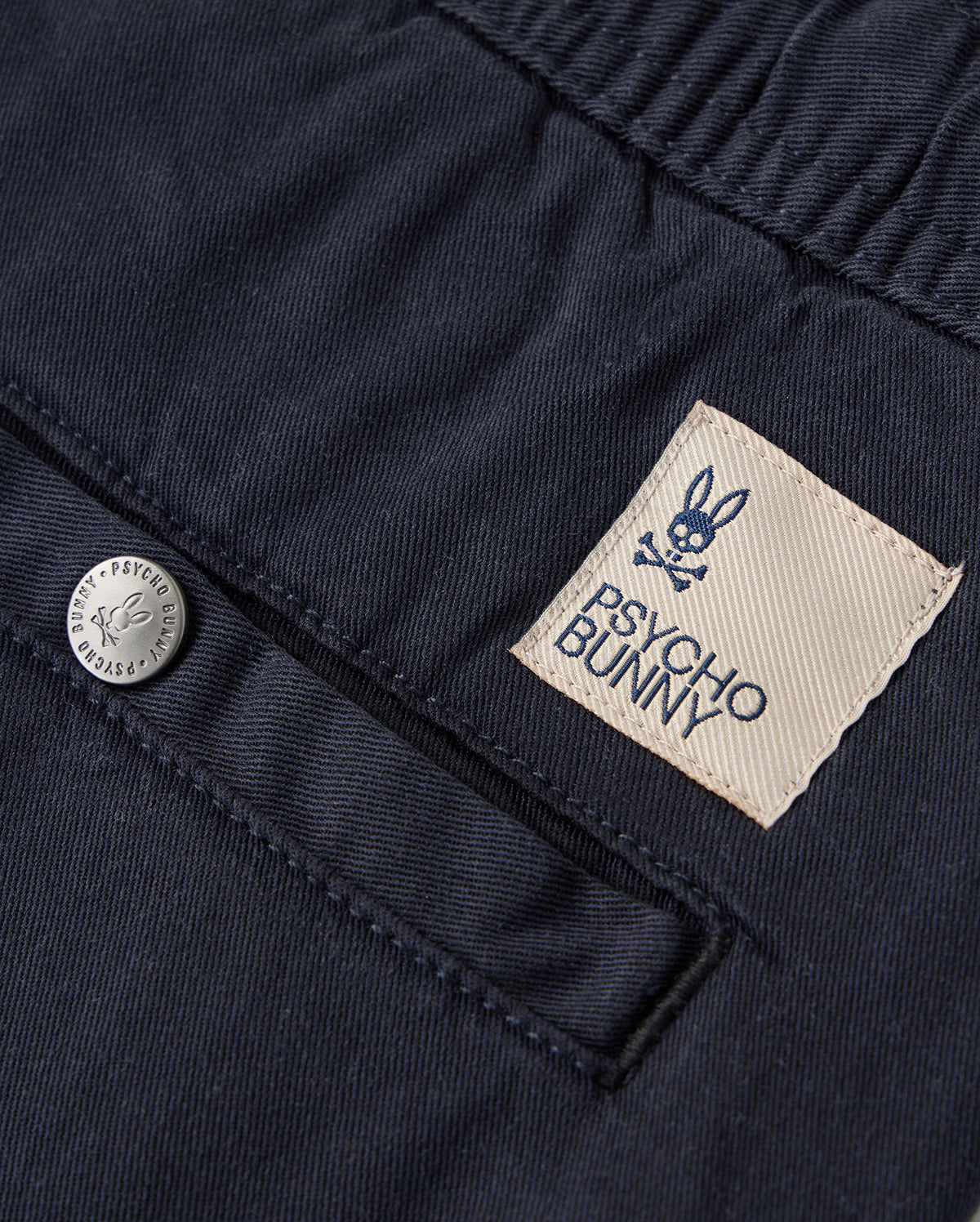 Close-up view of a navy blue KIDS WILLIS STRETCH TENCEL SHORT fabric with a textured pattern, featuring a beige label stitched with the "Psycho Bunny" logo and a metallic button engraved with "PSYCHO BUNNY PIMA COTTON.