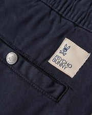 Close-up view of a navy blue KIDS WILLIS STRETCH TENCEL SHORT fabric with a textured pattern, featuring a beige label stitched with the "Psycho Bunny" logo and a metallic button engraved with "PSYCHO BUNNY PIMA COTTON.