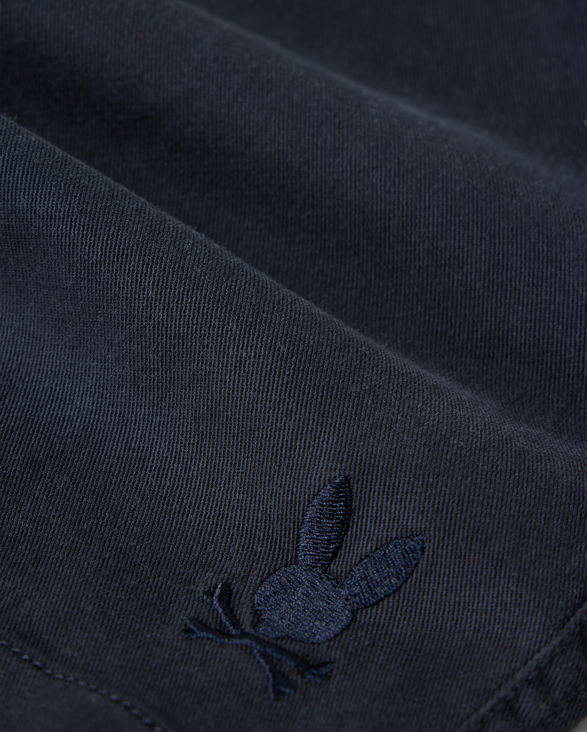 Close-up of Psycho Bunny kids' shorts made from dark KIDS WILLIS STRETCH TENCEL SHORT - B0R239Y1WB twill with a subtle, embroidered logo of a rabbit. The embroidery detail is in a slightly lighter shade of the same color as the fabric.