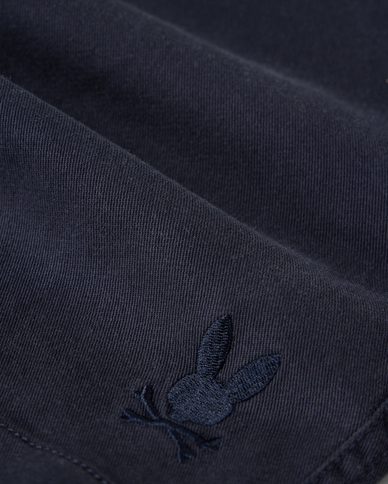 Close-up of Psycho Bunny kids' shorts made from dark KIDS WILLIS STRETCH TENCEL SHORT - B0R239Y1WB twill with a subtle, embroidered logo of a rabbit. The embroidery detail is in a slightly lighter shade of the same color as the fabric.