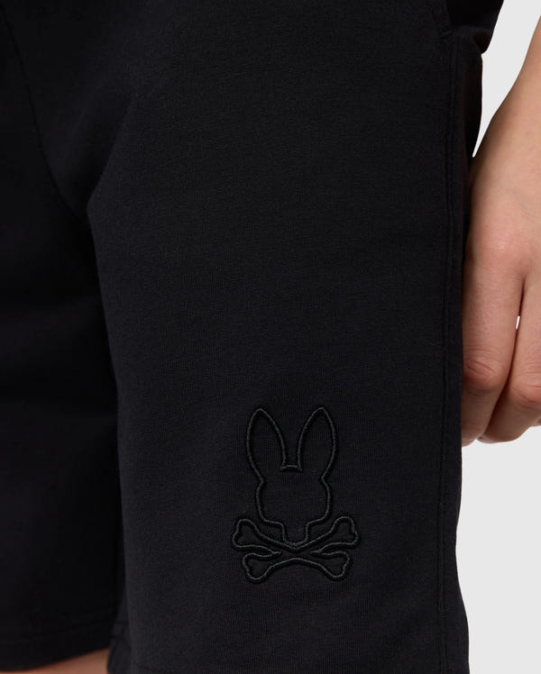Close-up image of a person wearing black Psycho Bunny KIDS LIVINGSTON TERRY SWEATSHORT - B0R408B200 with an embroidered logo on the leg. The logo features a silhouette of a rabbit's head above two crossed bones. The person's hand is partially visible in the shot, adding to the scene's comfort and casual vibe.