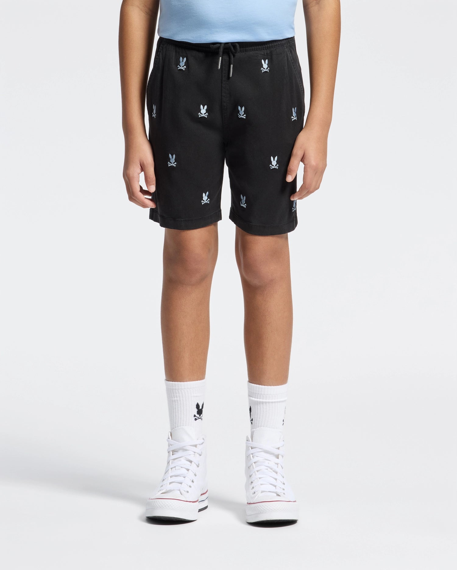 A person models Psycho Bunny's KIDS ALL OVER WILLIS STRETCH TENCEL SHORT (B0R559F200) in a light blue shirt, black shorts with white logos, and white knee-high socks featuring black logos. They sport chic white high-top sneakers against a simple white background.