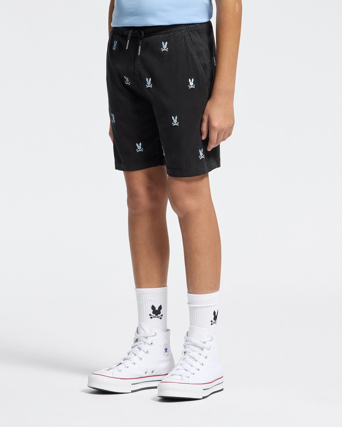 A person in Psycho Bunny's KIDS ALL OVER WILLIS STRETCH TENCEL SHORT - B0R559F200, featuring a black design with a bunny pattern, is paired with white high-top sneakers and socks. They're standing against a plain background, hands at their sides.