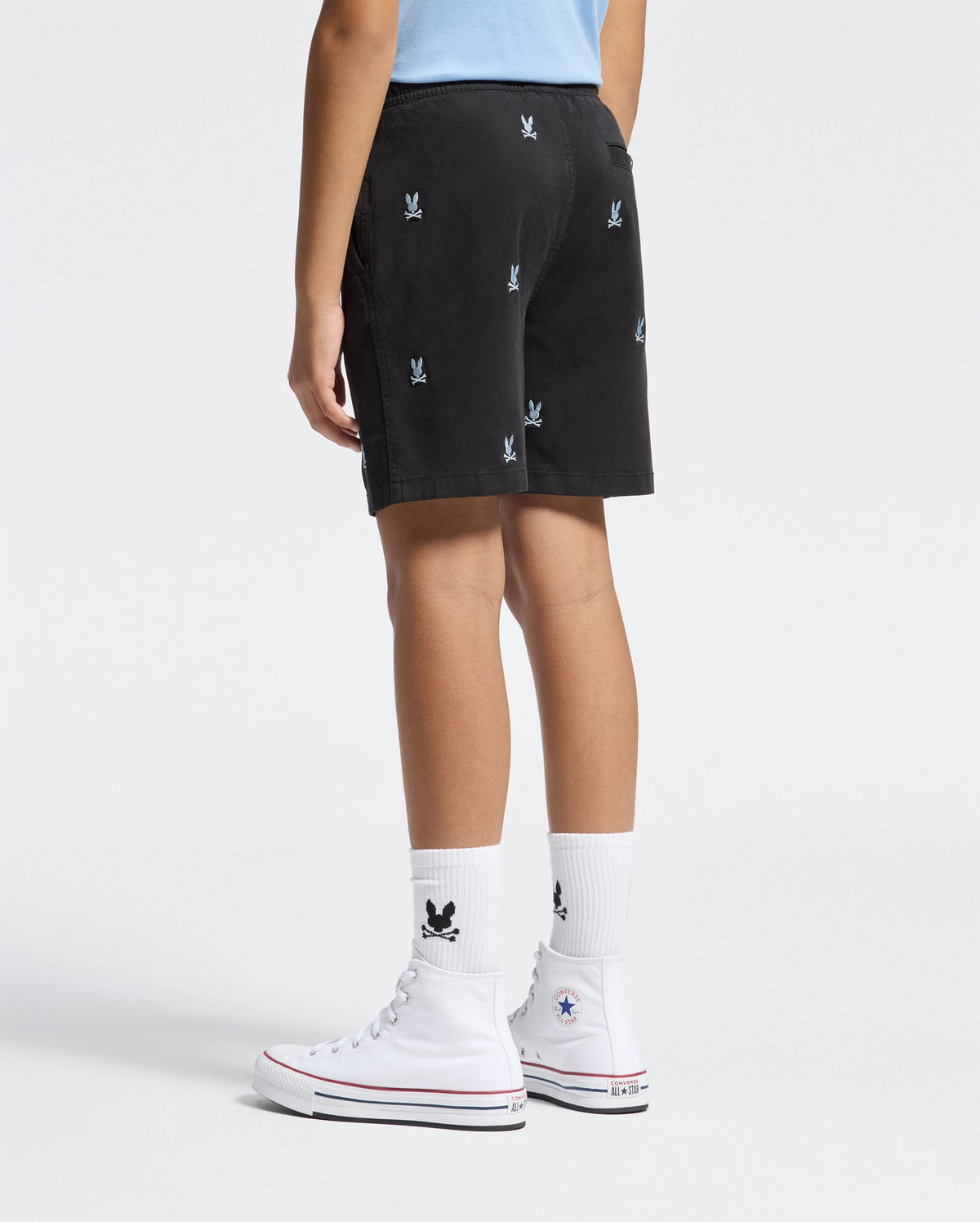 A person dressed in KIDS ALL OVER WILLIS STRETCH TENCEL SHORT by Psycho Bunny, featuring regular fit black shorts with small white logos, paired with white socks bearing a black logo and white high-top Converse sneakers against a plain white background.