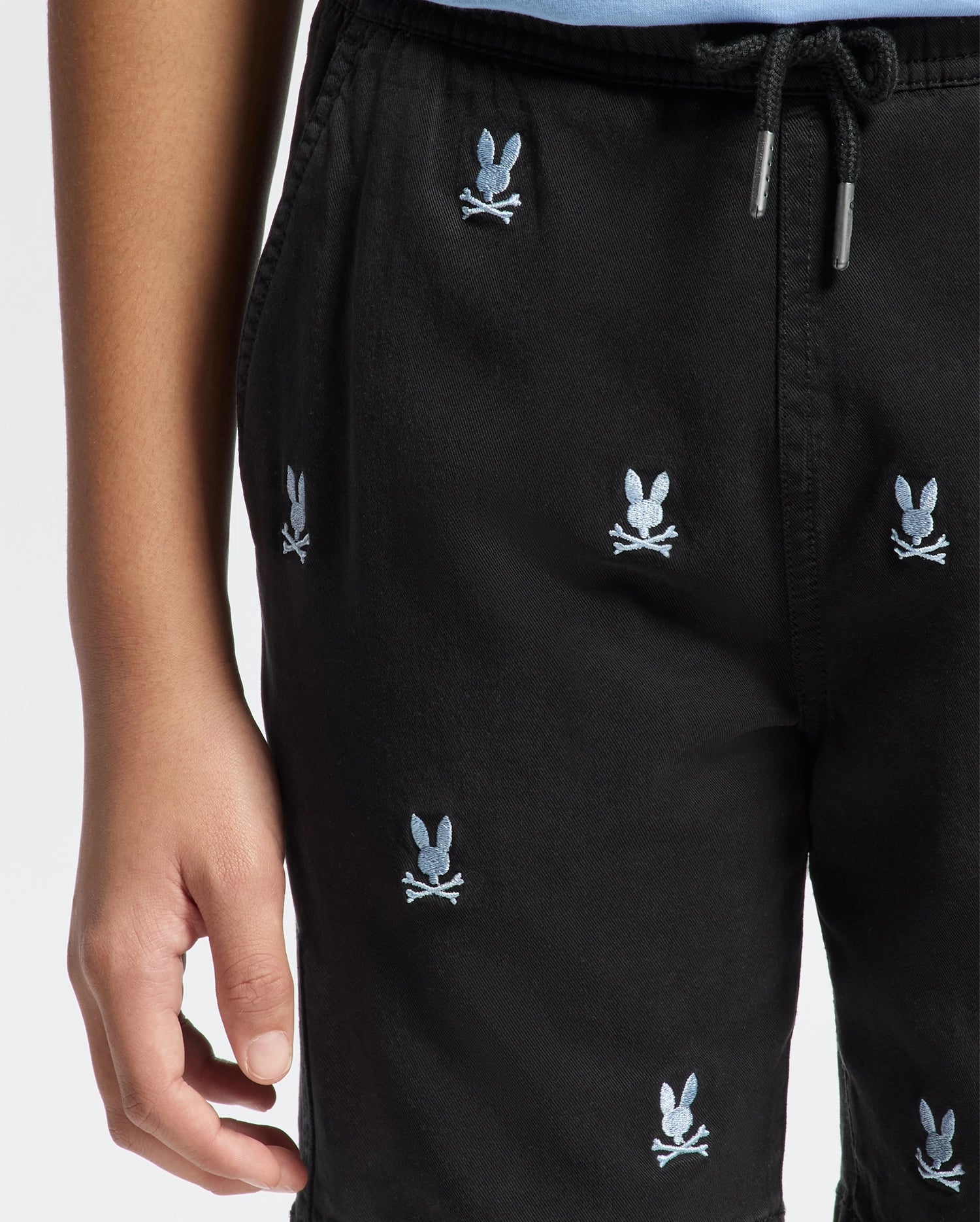 A person is wearing Psycho Bunny’s KIDS ALL OVER WILLIS STRETCH TENCEL SHORT (B0R559F200) with a black base featuring a light blue bunny and crossed bones pattern. These regular-fit shorts are made from Stretch Tencel™ and have a drawstring waist, set against a plain white background.