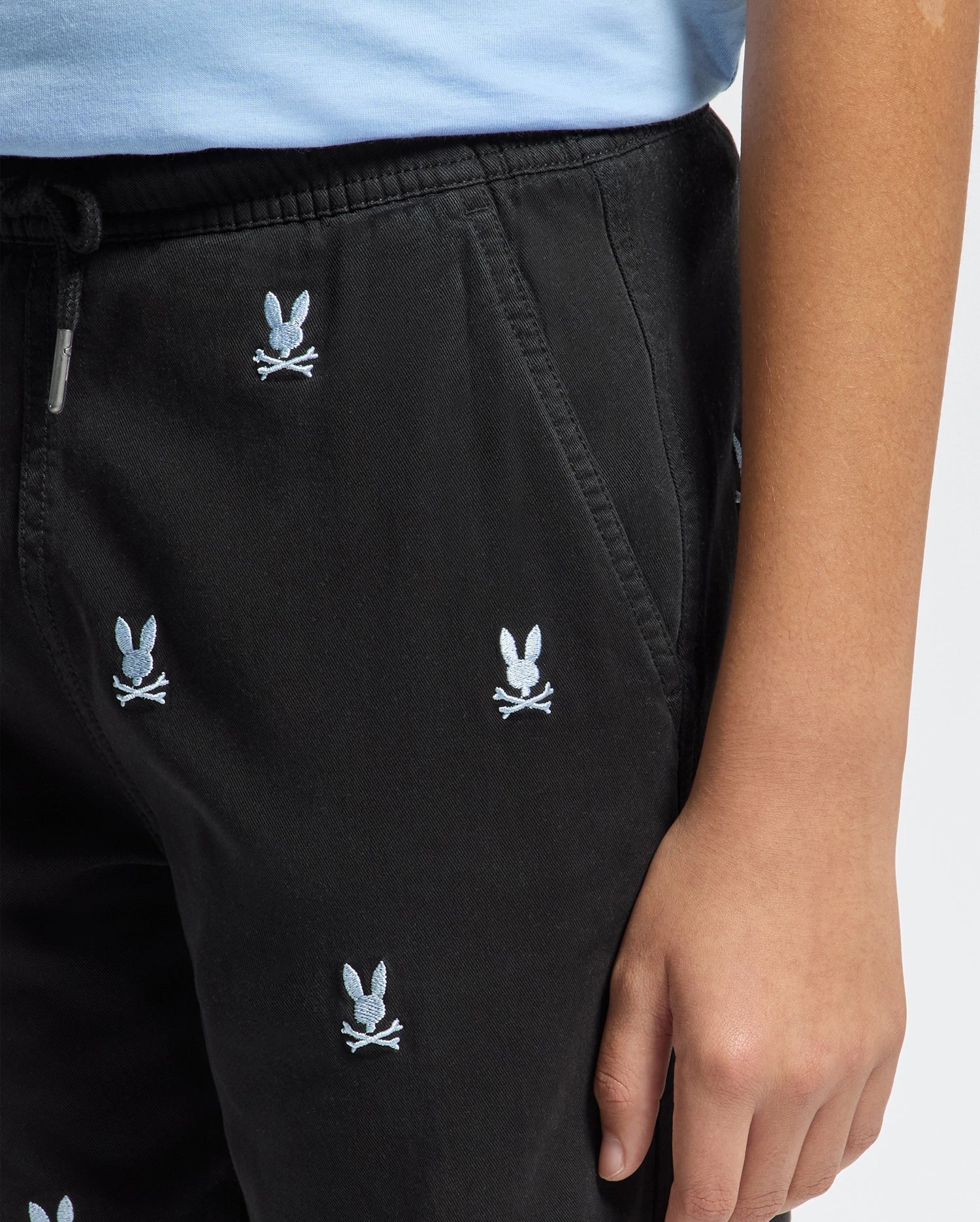 A person is dressed in black KIDS ALL OVER WILLIS STRETCH TENCEL SHORT by Psycho Bunny, featuring a blue embroidered bunny head and crossbones. Their right hand rests at their side, perfectly matching with a light blue top for a regular fit.