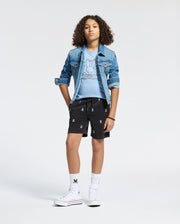 A young person poses against a light background wearing a denim jacket, a bunny-patterned light blue T-shirt from Psycho Bunny, black KIDS ALL OVER WILLIS STRETCH TENCEL SHORTS - B0R559F200 with white logos, white knee-high socks, and high-top sneakers. Their hands rest casually in the jacket pockets.