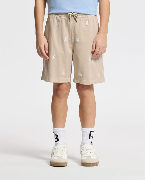 A person wears Psycho Bunny beige KIDS ALL OVER WILLIS STRETCH TENCEL SHORTS with a bunny pattern, hands in pockets; paired with a light blue shirt, white socks featuring black text, and white sneakers, against a plain white background.