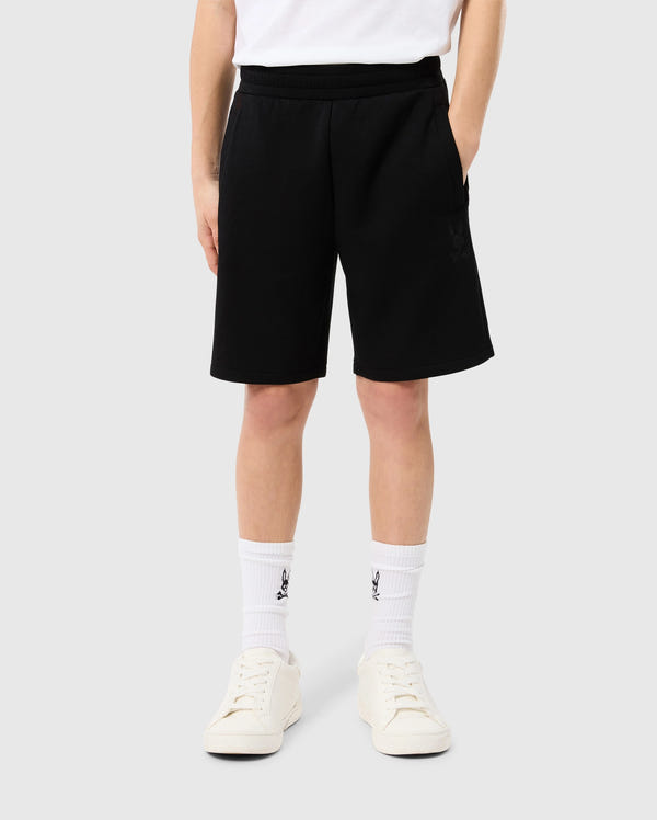 A person wearing Psycho Bunny KIDS LYONS SWEATSHORT - B0R578C200, white socks with a logo at the calf, and off-white sneakers, standing with their back to the camera. Only the lower half of the body is visible.